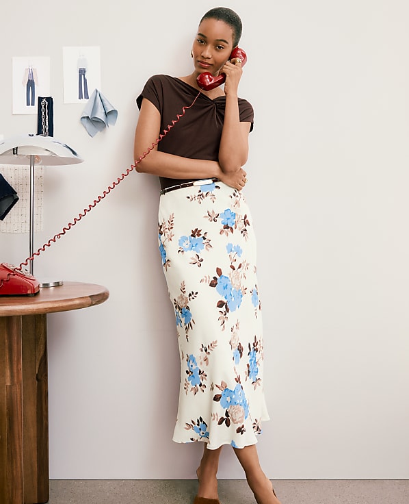 A woman in a floral skirt and brown top talks on a red rotary phone in front of a wall with sketches and a lamp on a desk.