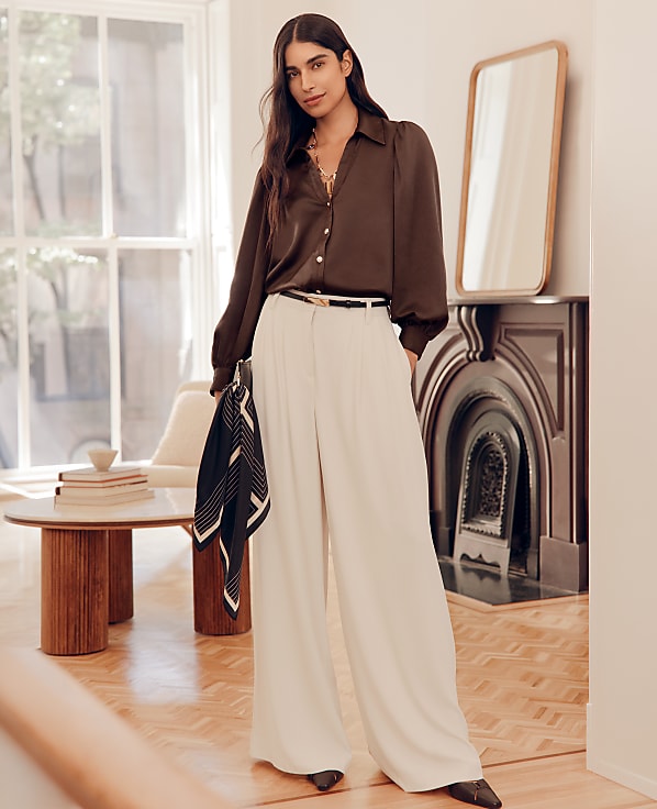 A woman in a brown blouse and cream wide-leg pants stands in a room with a fireplace and a round table.