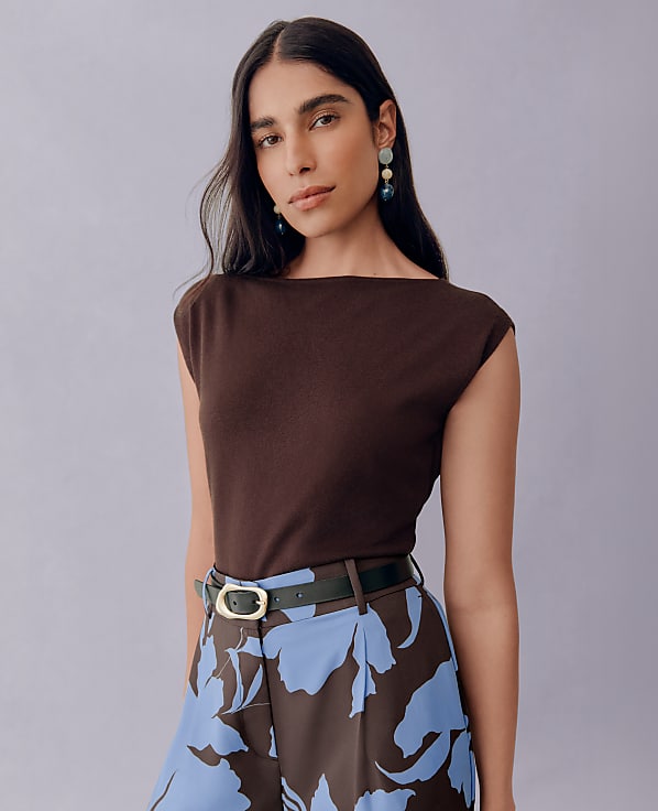 A woman with dark hair and large earrings stands against a plain purple background, wearing a brown top and patterned pants.