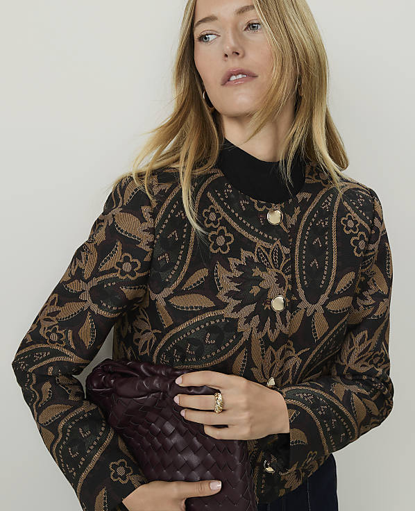 A blonde woman in a patterned jacket holds a woven clutch against a plain background.