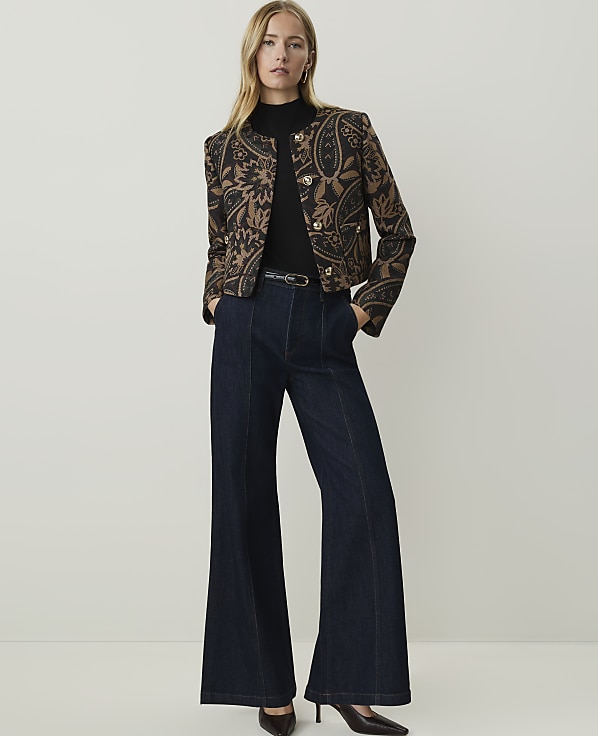 A woman in a patterned jacket and wide-leg jeans stands against a plain, light background.