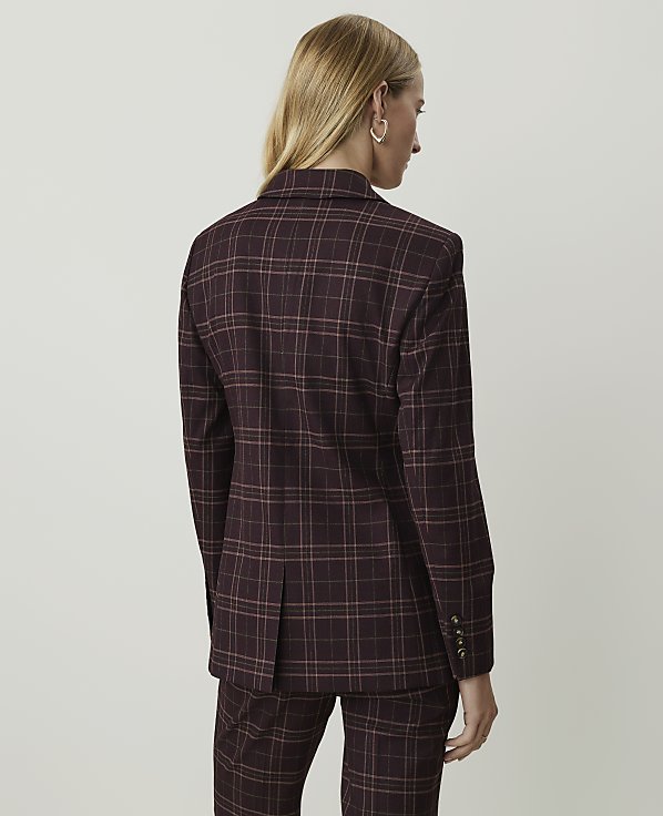 A woman in a burgundy plaid suit stands against a plain, light gray background.