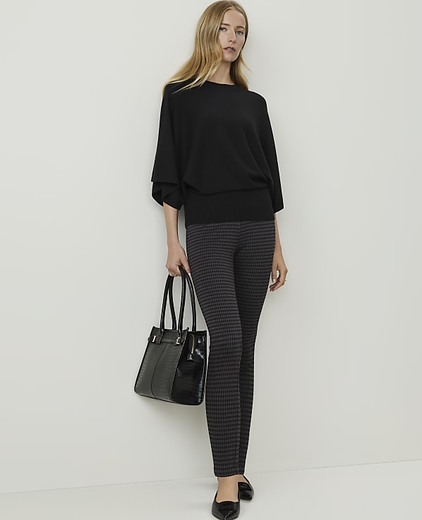 A woman in a black sweater and patterned pants stands with a black handbag against a plain white background.