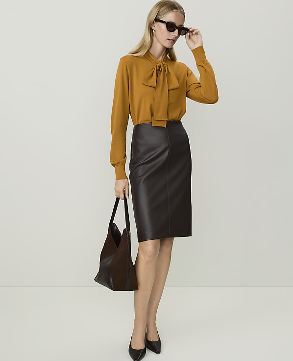 A woman in a mustard-colored bow-neck sweater and a dark brown leather pencil skirt stands with a dark handbag against a plain white background.