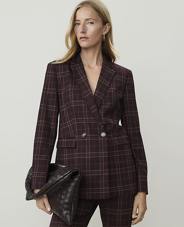 A woman in a burgundy plaid blazer and matching pants holds a woven clutch in front of a plain gray background.