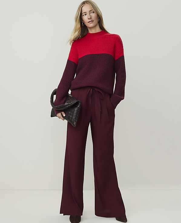 A woman in a two-toned red and burgundy sweater and matching wide-leg pants holds a dark clutch against a plain, light background.