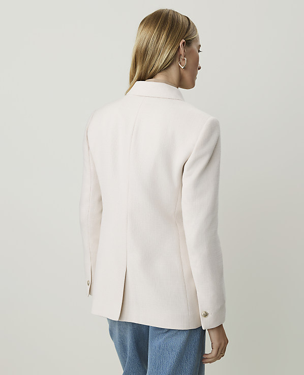 A woman in a light pink blazer and blue jeans stands against a plain, light-colored background.