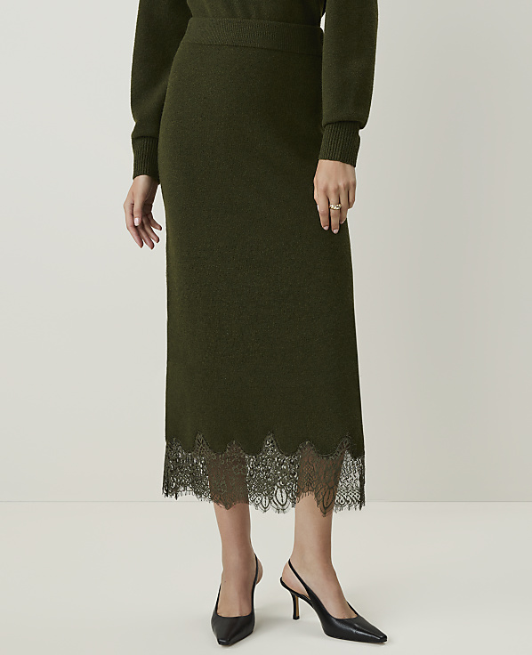 A person stands in a plain white studio, wearing a dark green knit midi skirt with a black lace hem and black slingback heels.