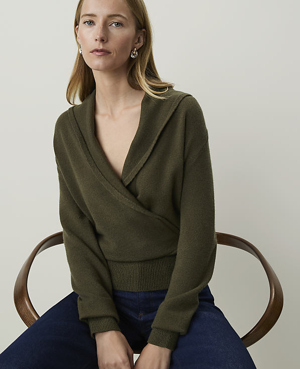 A woman in a green wrap sweater and blue jeans sits on a wooden chair against a plain background.
