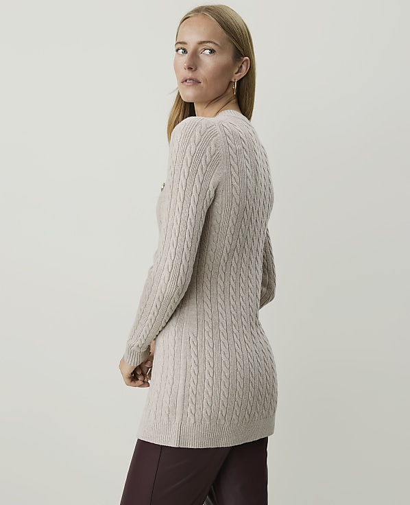 A woman in a beige cable-knit sweater and burgundy pants stands against a plain, light gray background.