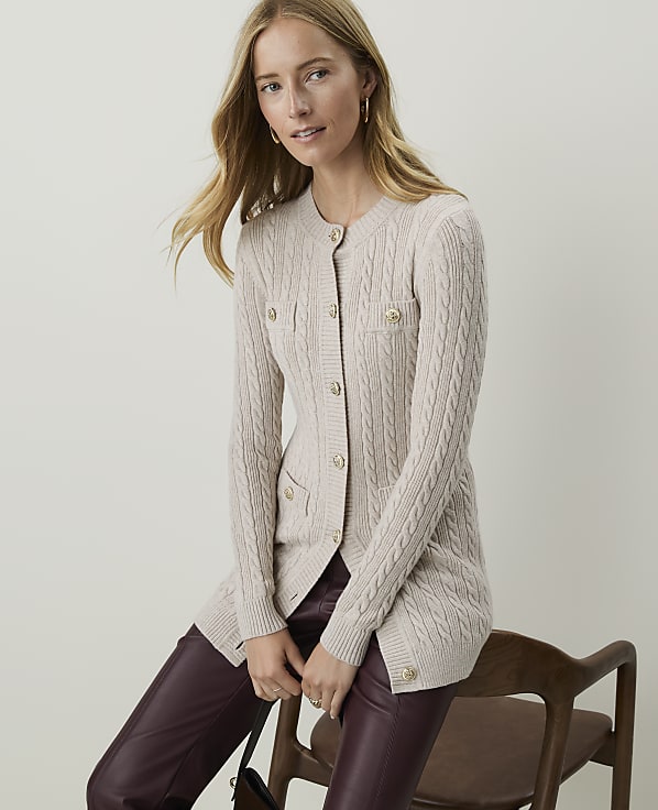 A woman in a cream cable-knit cardigan and burgundy pants sits on a wooden chair against a plain, light background.