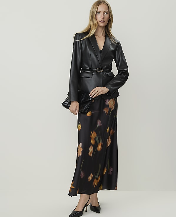 A woman in a black leather blazer and a floral print skirt stands against a plain white background.