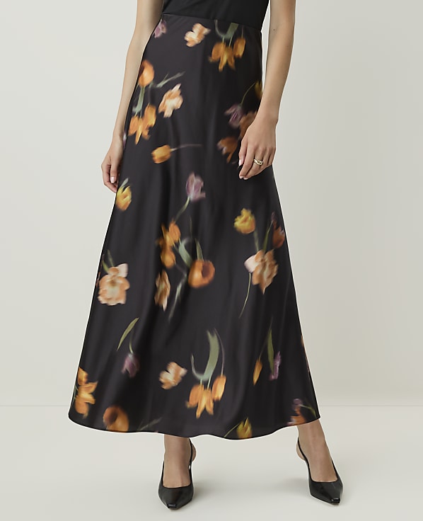A woman in a black floral skirt and heels stands against a plain, light-colored background.