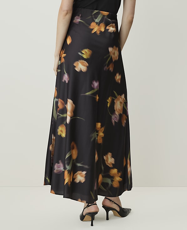 A person wearing a long, black floral skirt with orange and purple flowers stands against a plain, light-colored background.