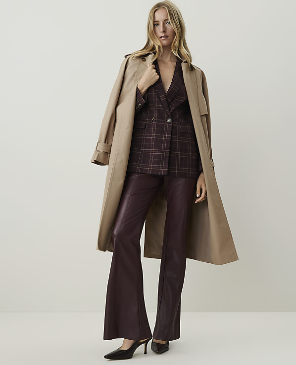 A woman in a plaid blazer and burgundy flared pants stands against a plain, light background, with a tan trench coat draped over her shoulders.