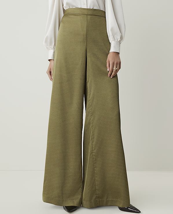 A person stands in wide-leg olive green patterned pants and a white blouse against a plain white background.