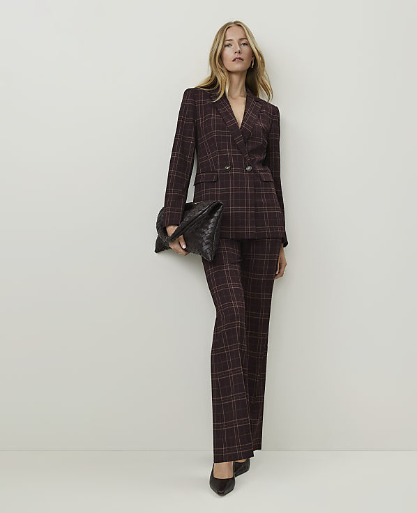 A woman in a plaid suit and holding a clutch stands against a plain, light-colored wall.