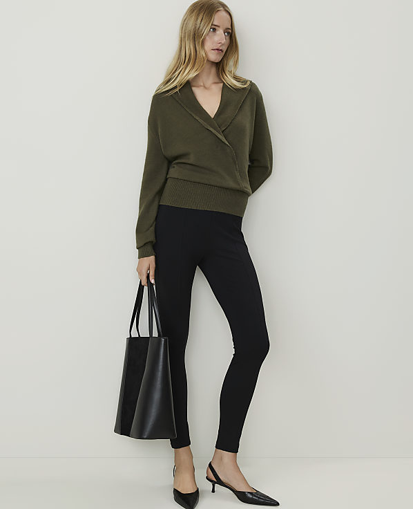 A woman in a green wrap sweater and black leggings stands with a black tote bag against a plain white background.
