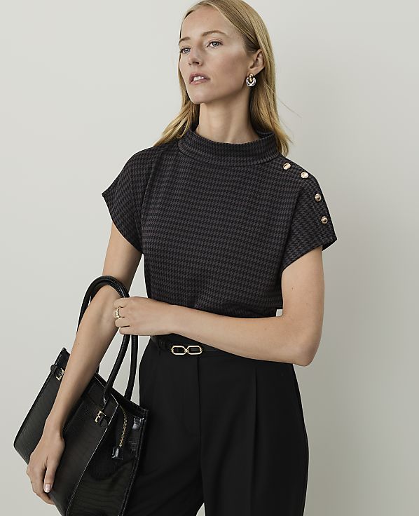 A woman in a patterned top and black pants holds a black handbag in front of a plain background.