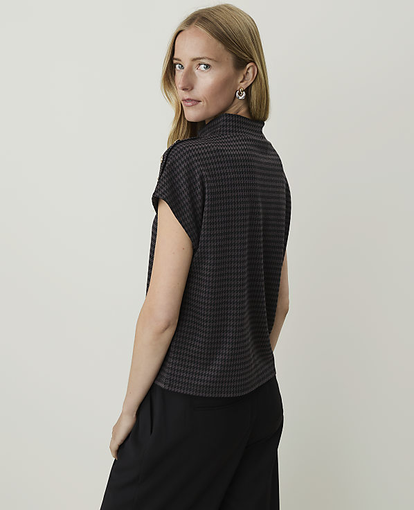 A woman with blonde hair and fair skin, wearing a dark patterned top and black pants, is shown from the back against a plain, light gray background.