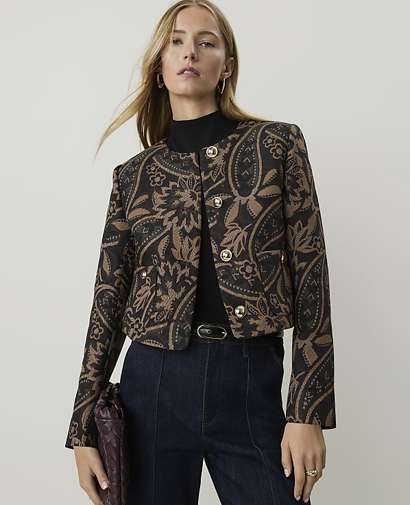A woman in a patterned jacket and dark jeans stands against a plain, light background.