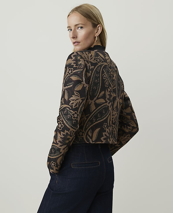 A woman in a patterned jacket and dark jeans stands against a plain background.