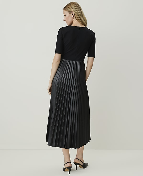 A woman in a black dress with a pleated faux leather skirt stands against a plain, light-colored background.