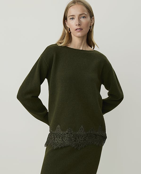 A woman in a green sweater with lace trim stands against a plain gray background.