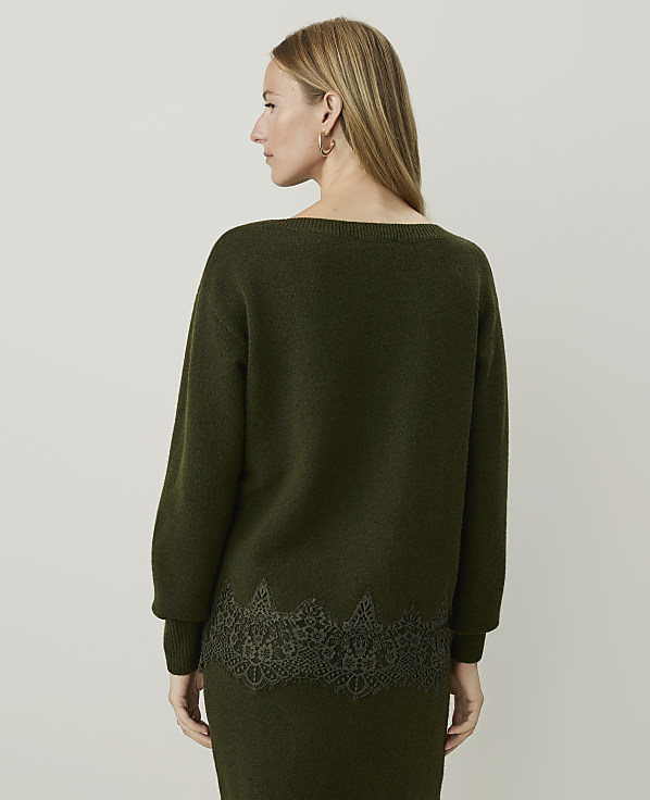 A woman in a green sweater with lace trim stands against a plain, light gray background.