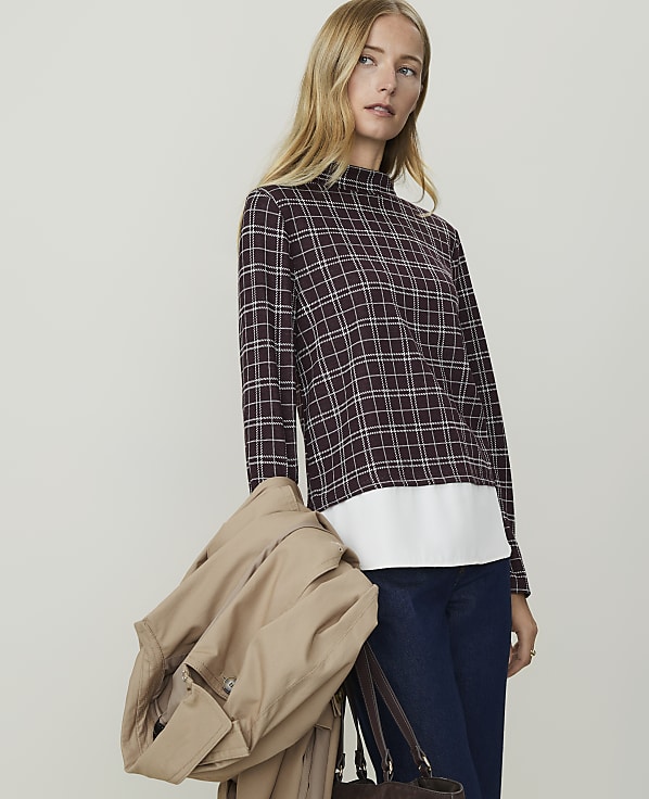 A woman in a plaid top and jeans carries a tan coat and a brown bag against a plain background.