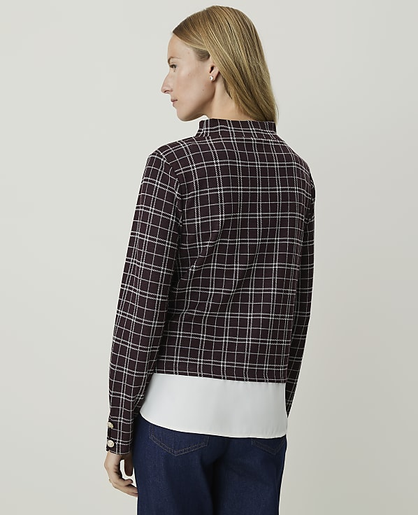 A woman in a plaid jacket and white shirt stands against a plain, light-colored background.