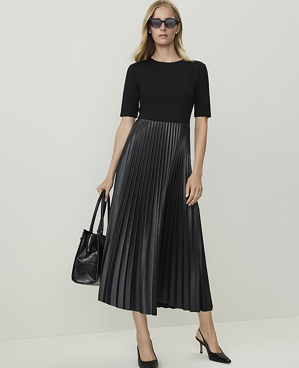 A woman in a black top and pleated faux leather skirt stands against a plain white background, holding a black handbag.