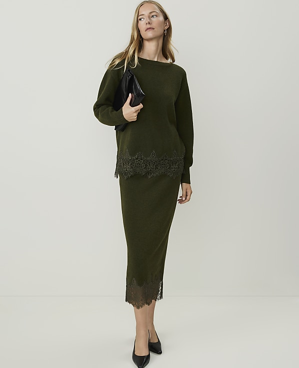 A woman in a green sweater and skirt with lace trim stands against a plain white background, holding a black clutch.
