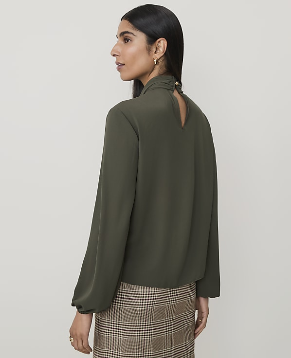 A woman in a green blouse and plaid skirt stands against a plain, light brown background.