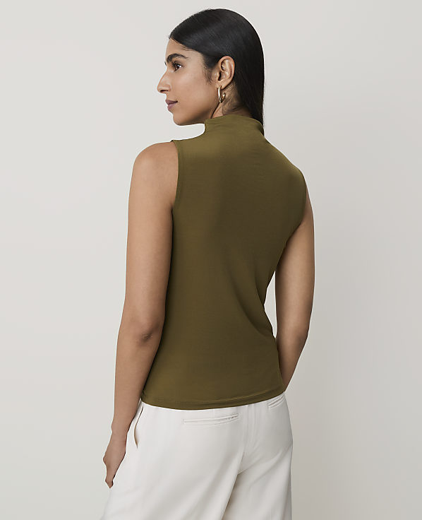 A woman in a sleeveless olive green turtleneck and white pants stands against a plain white background.