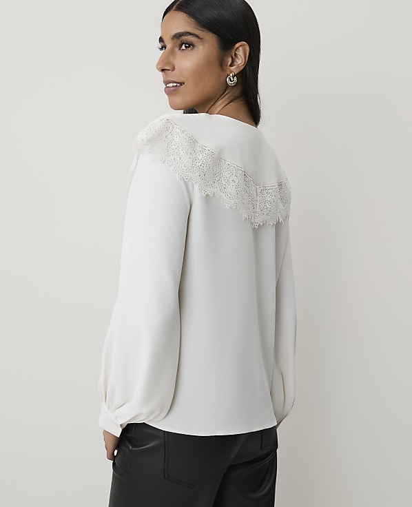 A woman in a white lace-trimmed blouse and black pants stands against a plain white background.
