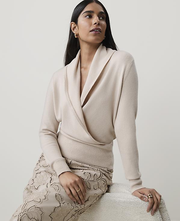A woman in a cream wrap sweater and patterned skirt sits against a plain, light background.