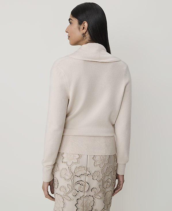 A woman in a cream cowl-neck sweater and a patterned skirt stands against a plain, light background.
