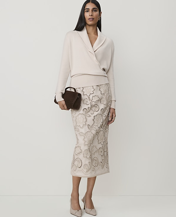 A woman in a cream wrap sweater and a beige embroidered skirt stands against a plain white background, holding a small brown clutch.