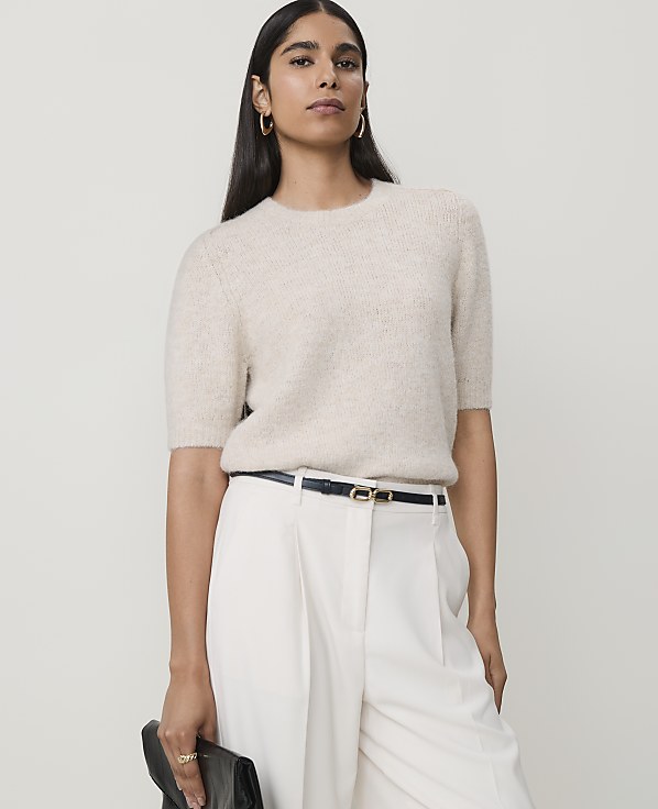 A woman in a cream sweater and white pants stands against a plain, light background, holding a black clutch.