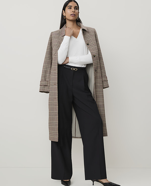 A woman in a white top, black wide-leg pants, and a plaid coat stands against a plain, light-colored background.