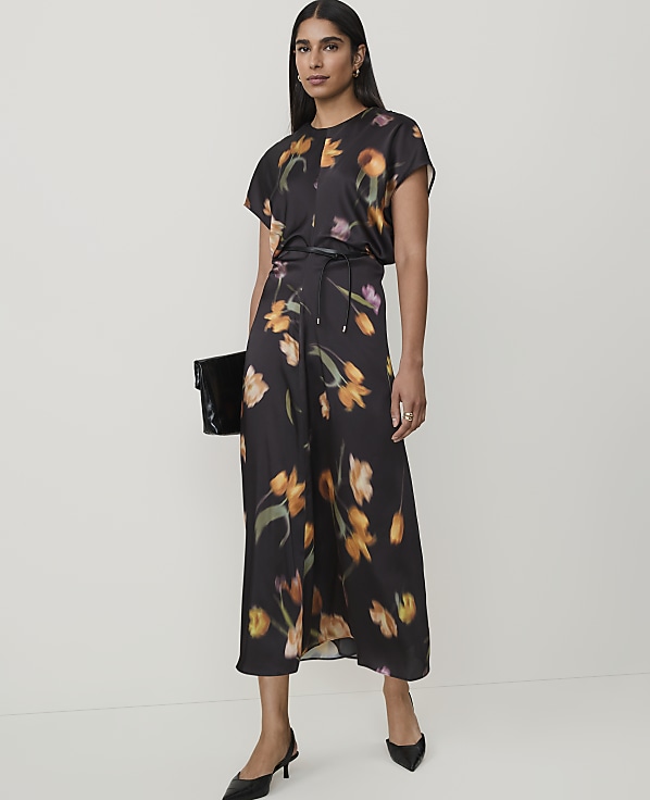 A woman in a floral dress and black heels stands against a plain background, holding a black clutch.