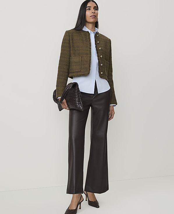 A woman in a tweed jacket and leather pants stands against a plain white background, holding a woven clutch.