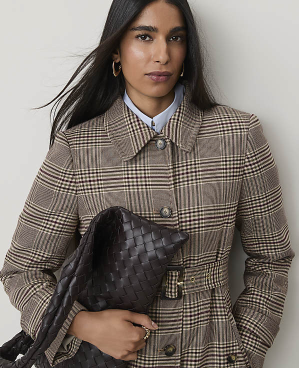A woman in a plaid coat holds a woven bag in front of a plain background.