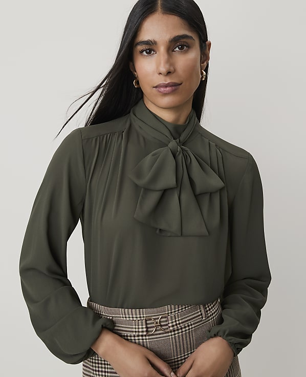 A woman in a green bow-neck blouse and plaid skirt stands against a plain, light background.