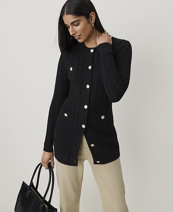 A woman in a black cable-knit cardigan with gold buttons holds a black tote bag in front of a plain background.