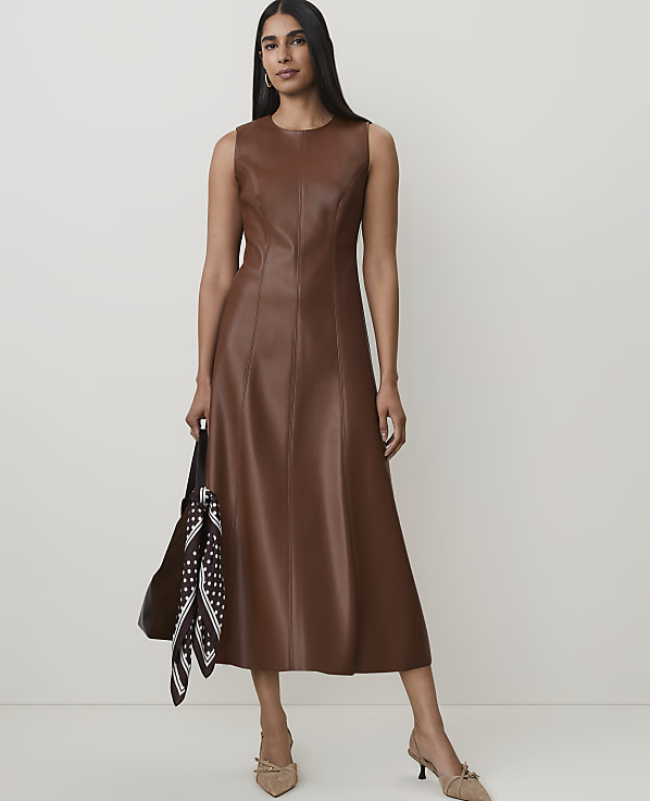 A woman in a brown leather dress and heels stands against a plain, light background, holding a black bag with a patterned scarf.