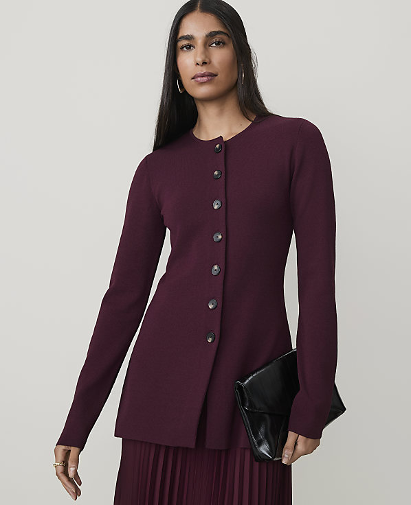 A woman in a burgundy cardigan and pleated skirt holds a black clutch against a plain background.