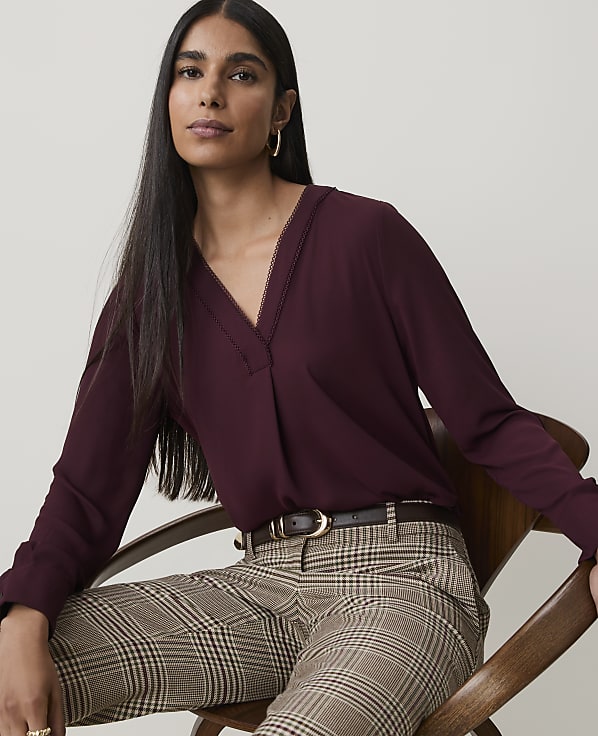 A woman in a burgundy blouse and plaid pants sits in a chair against a plain background.