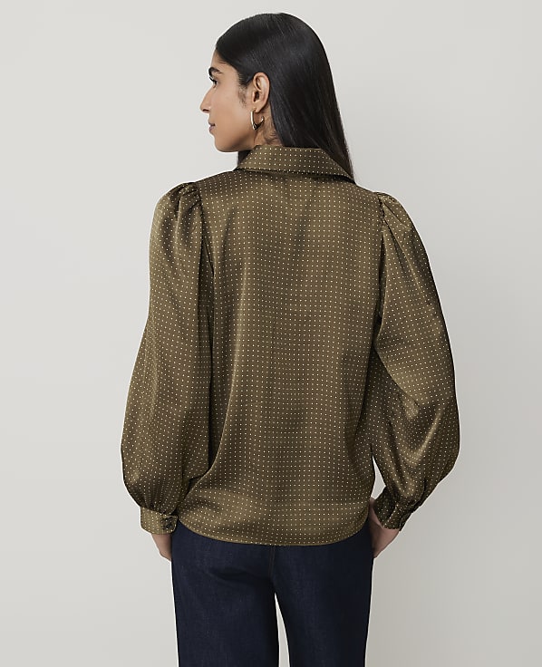 A woman in a patterned olive green blouse with puffed sleeves stands against a plain, light brown background.
